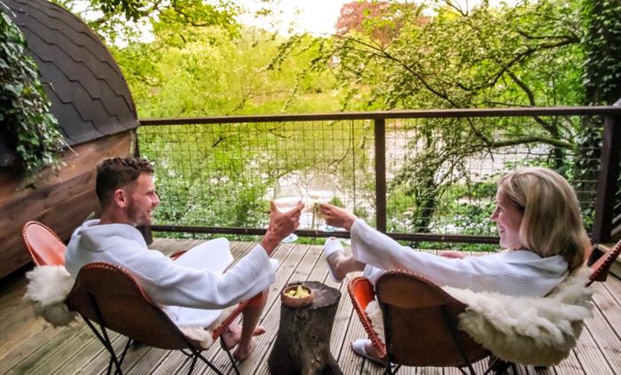 A couple raises glasses in a toast, sat in robes in comfy chairs on decking overlooking the river.
